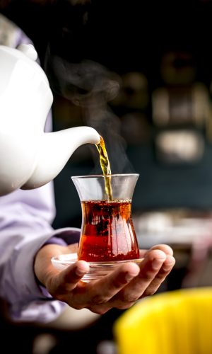 side view of a person pouring black tea from white ceramic teapot into armudu glass